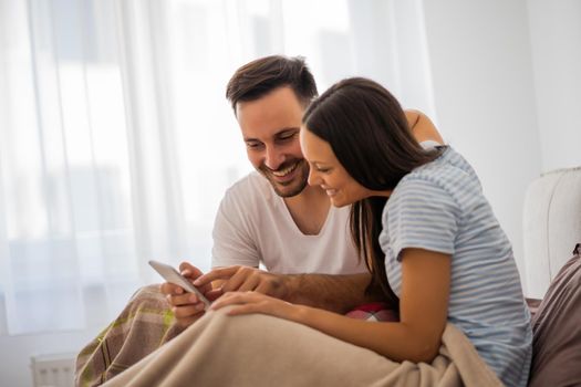 Young happy couple is looking at smartphone in bed in the morning.