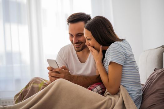 Young happy couple is looking at smartphone in bed in the morning.