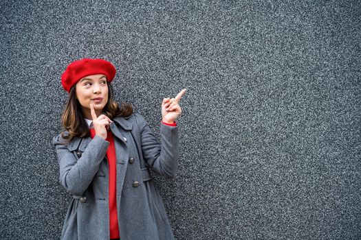 Adult woman in modern casual clothing standing in front of gray background. She is pointing at copy space for your advert.