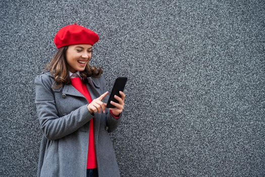 Adult woman in modern casual clothing standing in front of gray background. She is messaging on phone. Copy space for your advert.