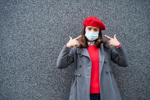 Adult woman in modern casual clothing standing in front of gray background. She is pointing at protective mask. Copy space for your advert.