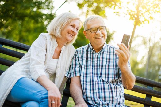 Happy senior couple is using smartphone in park. They are taking selfie.