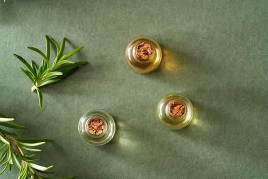 Three bottles of essential oil with fresh rosemary twigs on green paper background, top view