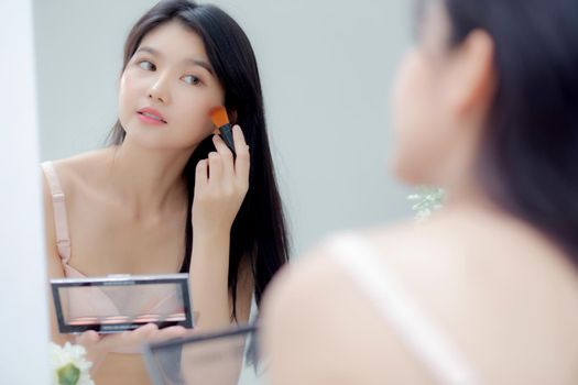 Beauty young asian woman happy and smile with face looking mirror applying makeup with brusher of cheek in the room, beautiful of girl holding brush make-up on facial, skin care and cosmetic concept.