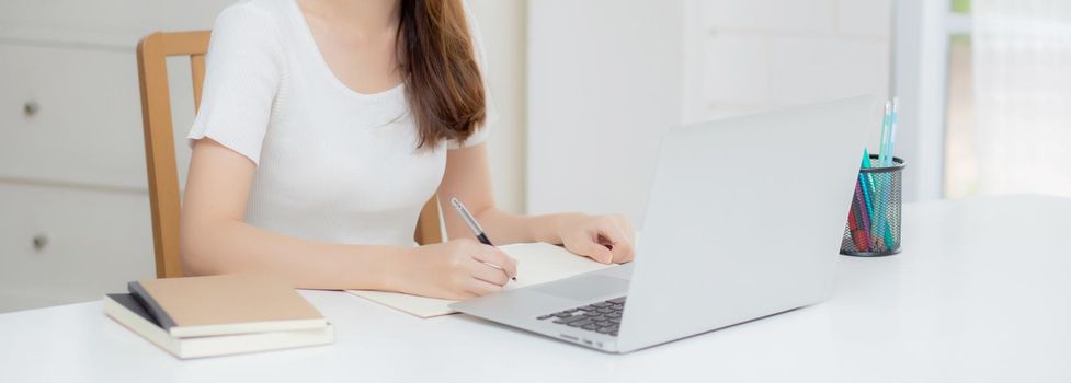 Young asian woman writing on notebook for planning working and using laptop computer on desk at home, girl notes about finance, female study and learning, business and communication concept.