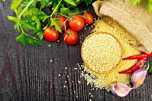 Raw couscous in a bowl and a sack of burlap, garlic, tomatoes, hot peppers and herbs on black wooden board background from above