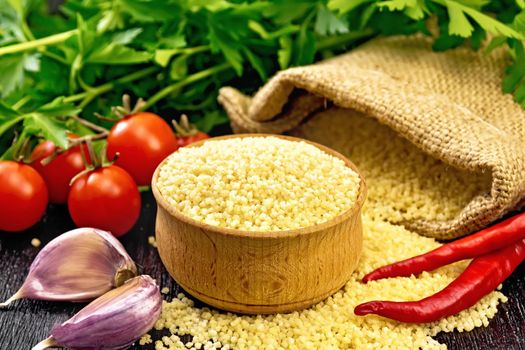 Raw couscous in a bowl and a sack of burlap, garlic, tomatoes, hot peppers and herbs on black wooden board background