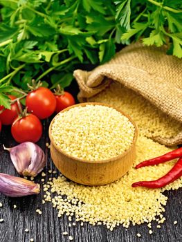Raw couscous in a bowl and a sack of burlap, garlic, tomatoes, hot peppers and herbs on the background of dark wooden board