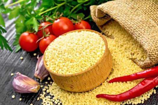 Raw couscous in a bowl and a sack of burlap, garlic, tomatoes, hot peppers and herbs on wooden board background