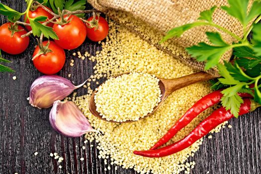 Raw couscous in a spoon and a sack of burlap, tomatoes, hot peppers, herbs and garlic on wooden board background from above