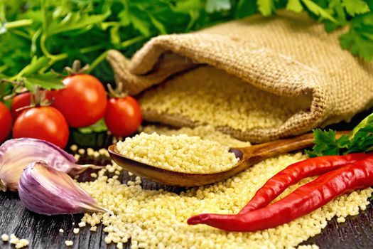 Raw couscous in a spoon and a sack of burlap, tomatoes, hot peppers, herbs and garlic on the background of dark wooden board