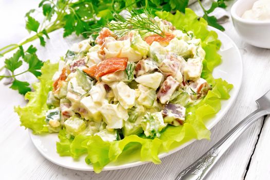 Salad from salmon, cucumber, eggs and avocado, dressed with mayonnaise on lettuce leaves in a plate, kitchen towel, dill, parsley and fork on a light wooden board background