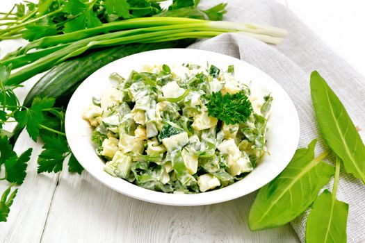 Salad of cucumber, sorrel, boiled potatoes, eggs and herbs, dressed with mayonnaise in a white plate, parsley, green onions and towel against the background of light wooden boards