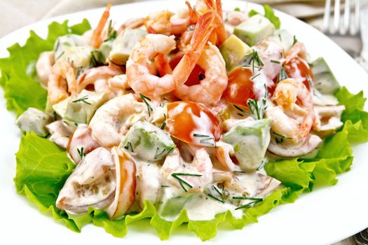 Salad with shrimp, avocado, tomatoes and mayonnaise on the lettuce in the plate, napkin, fork on the background light wooden board