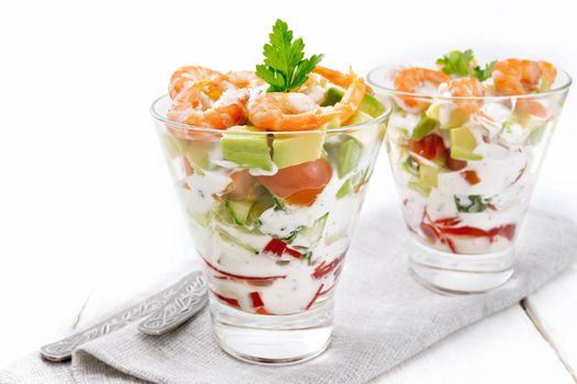 Puff salad with shrimp, avocado, fresh cucumber, sweet pepper and tomato, seasoned with yogurt sauce in two glass glasses on a napkin, bread and forks on a white wooden board background