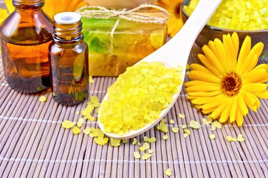 Bath salt in a spoon, two bars of homemade soap, oil in two bottles, calendula, a wooden mortar in the background of a bamboo napkin