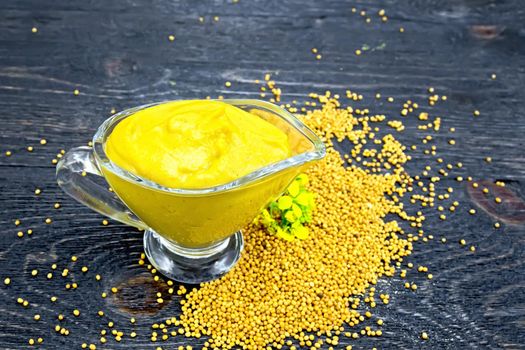 Mustard sauce in a glass sauceboat, seeds and flowers of mustard on a background of a dark wooden board