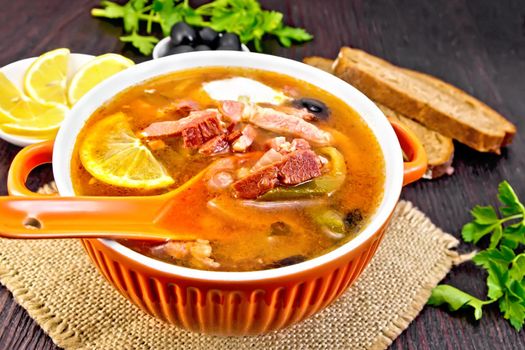 Soup saltwort with lemon, meat, pickles, tomato sauce olives, spoon in a bowl on a sacking, bread on a wooden board background