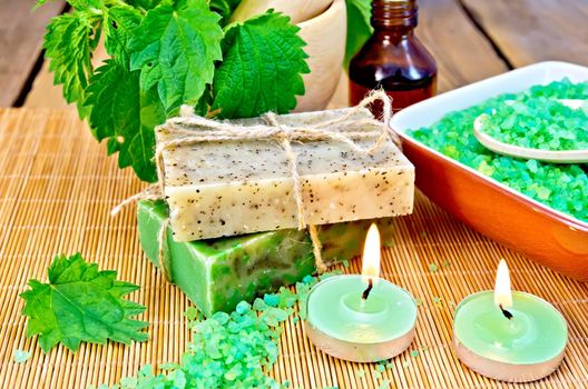 Two bars of homemade soap with twine, bath salt in a bowl, oil in the bottle, nettles in a mortar, two candles on a wooden boards background