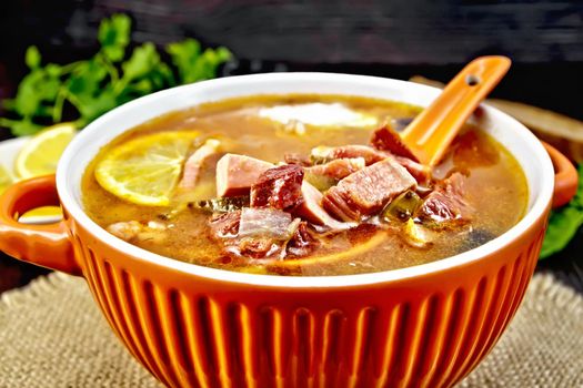 Soup saltwort with lemon, meat, pickles, tomato sauce olives, spoon in a bowl on a sacking, bread on a background of a dark wooden board