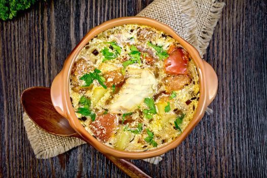 Fish soup with tomatoes, potatoes, peppers and cream in a clay bowl on a sackcloth napkin, parsley on a wooden board background from above