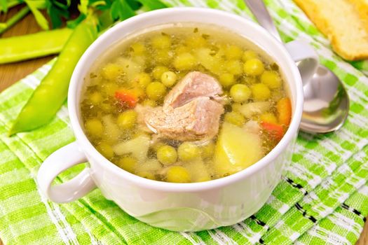 Soup of green peas with meat and potatoes in a white bowl, bread, spoon, pods on green napkin at the background of the wooden planks