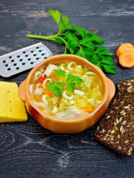 Minestrone soup with meat, celery, tomatoes, zucchini and cabbage, green peas, carrots and pasta in an earthenware bowl, cheese, grater on black background wooden plank