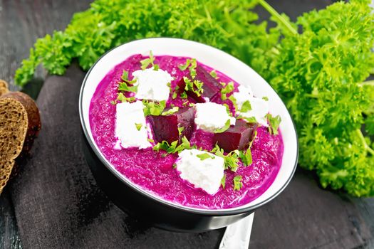 Soup puree of beetroot, potato and cream with salted cream cheese in a bowl on towel, bread and parsley on dark wooden board background