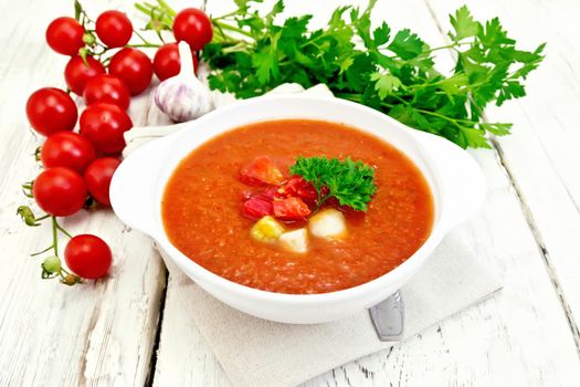 Gazpacho tomato soup in a white bowl with parsley and vegetables, spoon on a napkin on the background light wooden boards