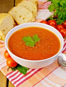 Tomato soup in a white bowl on a napkin with a spoon, tomato, parsley, bread on a wooden boards background