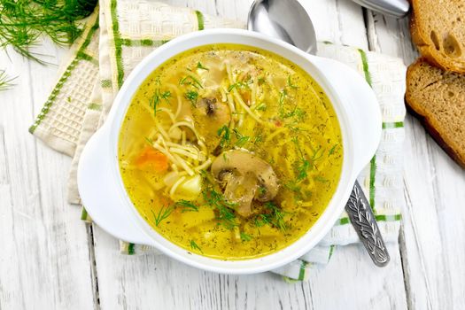 Soup with noodles and champignons in a white bowl on a napkin, spoon and bread on the background light wooden boards on top