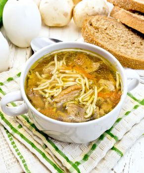Soup with noodles and mushrooms in a white bowl, bread, napkin and spoon on a background of bright wooden planks