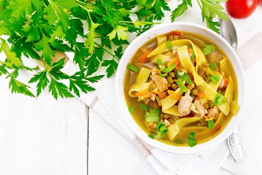 Soup with meat, tomatoes, vegetables, mung bean lentils and noodles in a bowl on napkin, parsley and a spoon on the background of light wooden board from above