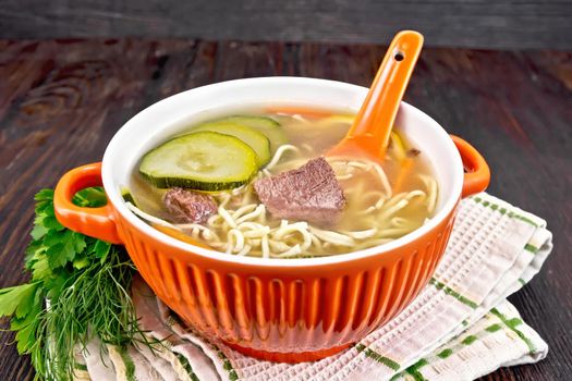 Soup with zucchini, beef, ham, lemon and noodles in a red bowl, parsley and dill on a towel on a wooden plank background
