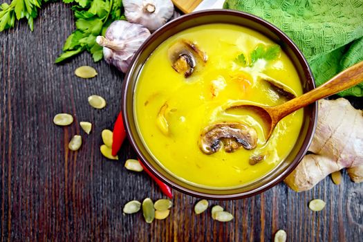 Soup-puree pumpkin with prawns and champignons, a spoon in a brown bowl, napkin, parsley, pumpkin seeds, ginger on a wooden board background