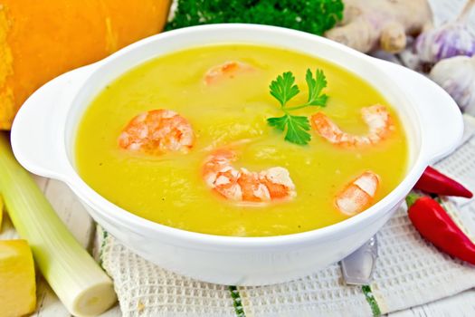 Soup-puree pumpkin with shrimp and parsley in a white bowl on a kitchen towel with a spoon, squash, parsley, ginger, hot red pepper pods, leek on a wooden boards background