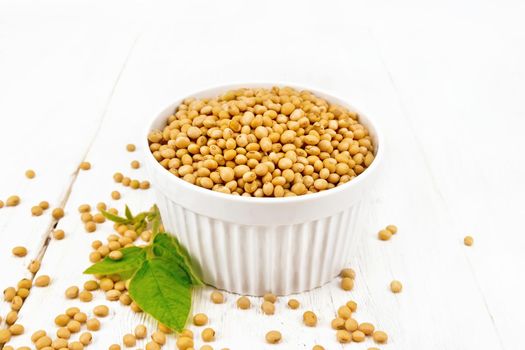 Soy beans in a bowl and on a table, green leaf on background of light wooden board