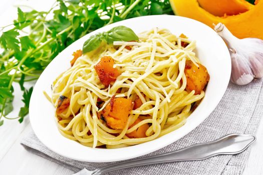 Spaghetti with pumpkin, onion and garlic in a plate on a napkin, basil, parsley and fork on white wooden board background