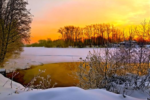 Water and ice on the river, shrubs and trees, wooden house on a background of sunset sky