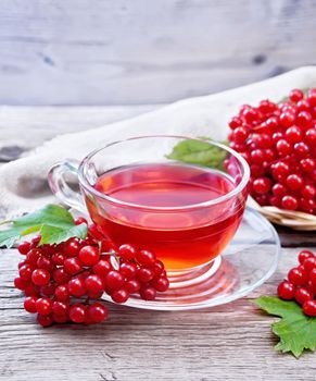 Tea with viburnum in a glass cup, wicker plate with berries, green leaves and burlap on wooden board background