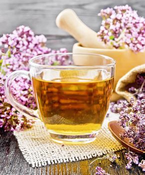 Oregano herbal tea in a glass cup on burlap, fresh flowers in mortar and on the table, dried marjoram flowers in a bag and spoon on wooden board background