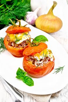 Tomatoes stuffed with meat and rice with cheese in a plate on a napkin, fork, dill and parsley, onion and basil on a wooden board background