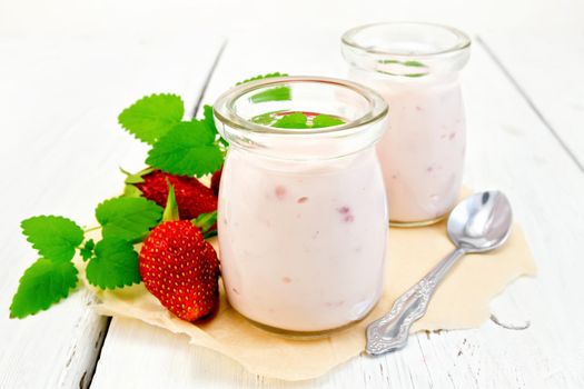 Yogurt with strawberries in two jars and a spoon, strawberries and mint on parchment on the background light wooden boards