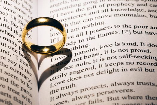 One golden wedding ring on Holy bible book with heart shaped shadow close up