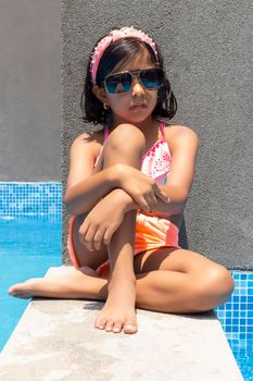 Cute latin girl is sitting on the edge of the pool, receiving the summer sun