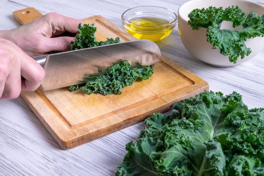 A female hand is cutting the leaves of kale with a large kitchen knife on a wooden cutting board. Super food concept, healthy and vegetarian food. Selective focus.