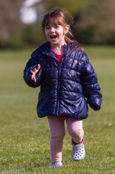 Full-length shot of a cute brown hair baby girl with a blue jacket and pink trousers running on the grass in a park on a sunny spring day