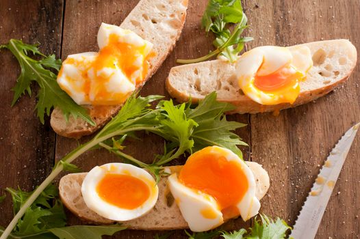 Soft boiled eggs broken open on sliced rolls or baguettes with fresh salad green and a knife while preparing a healthy sandwich