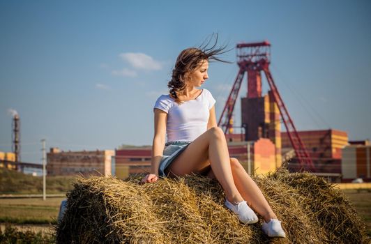 A young woman sits on a round bale of straw in a field against the background of a potash factory