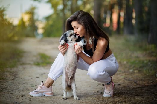 A young woman with her cute Shih Tzu is sitting on a path in the woods.